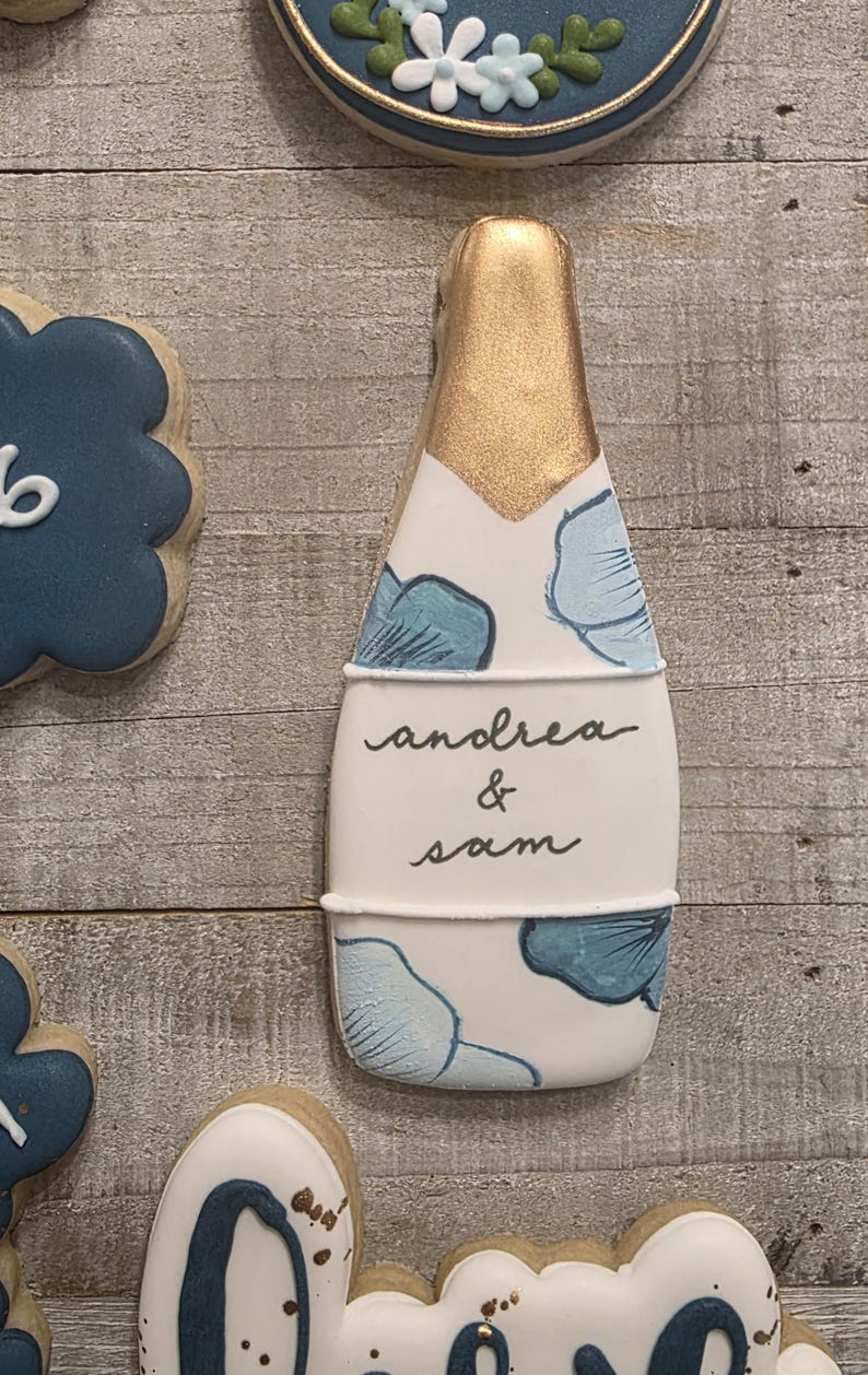 K&ouml;nnte beinhalten: Ein Keks in Champagnerflaschenform mit goldener Folienoberseite und blauem Blumendesign. Auf dem Keks stehen die Namen "andrea & sam". Weitere Kekse mit blau-wei&szlig;en Designs befinden sich im Hintergrund.