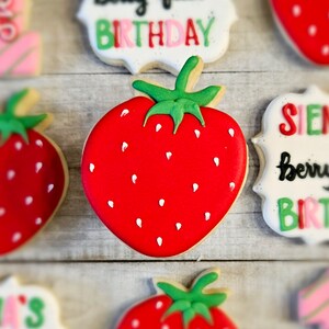 May include: A red and white decorated sugar cookie shaped like a strawberry with a green stem and leaves. The cookie is decorated with white dots to resemble seeds.