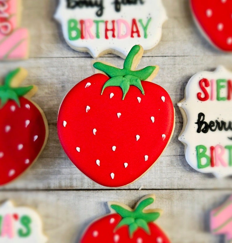 May include: A red and white decorated sugar cookie shaped like a strawberry with a green stem and leaves. The cookie is decorated with white dots to resemble seeds.