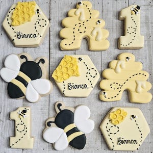 May include: Nine decorated sugar cookies with a bee theme. The cookies are yellow and feature bees, honeycombs, and the number one. Some cookies have the name Bianca written on them.
