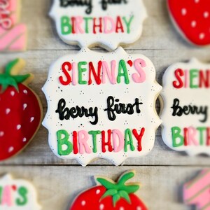 May include: A white cookie with a scalloped edge decorated with red, green, and pink icing. The cookie says "Sienna's berry first birthday" in black and white icing.