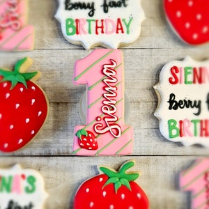 May include: A set of decorated sugar cookies with a strawberry theme. The cookies feature a pink and green number one with the name Sienna written on it, a cookie with the words "berry first birthday", a cookie with the words "one's first", and two cookies with strawberries.