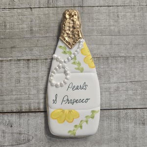 K&ouml;nnte beinhalten: Ein Champagnerflaschen-f&ouml;rmiger Keks mit goldenem Folienaufsatz, verziert mit einer Perlenkette und gelben Blumenakzenten. Die Worte "Pearls & Prosecco" sind auf der Vorderseite geschrieben.