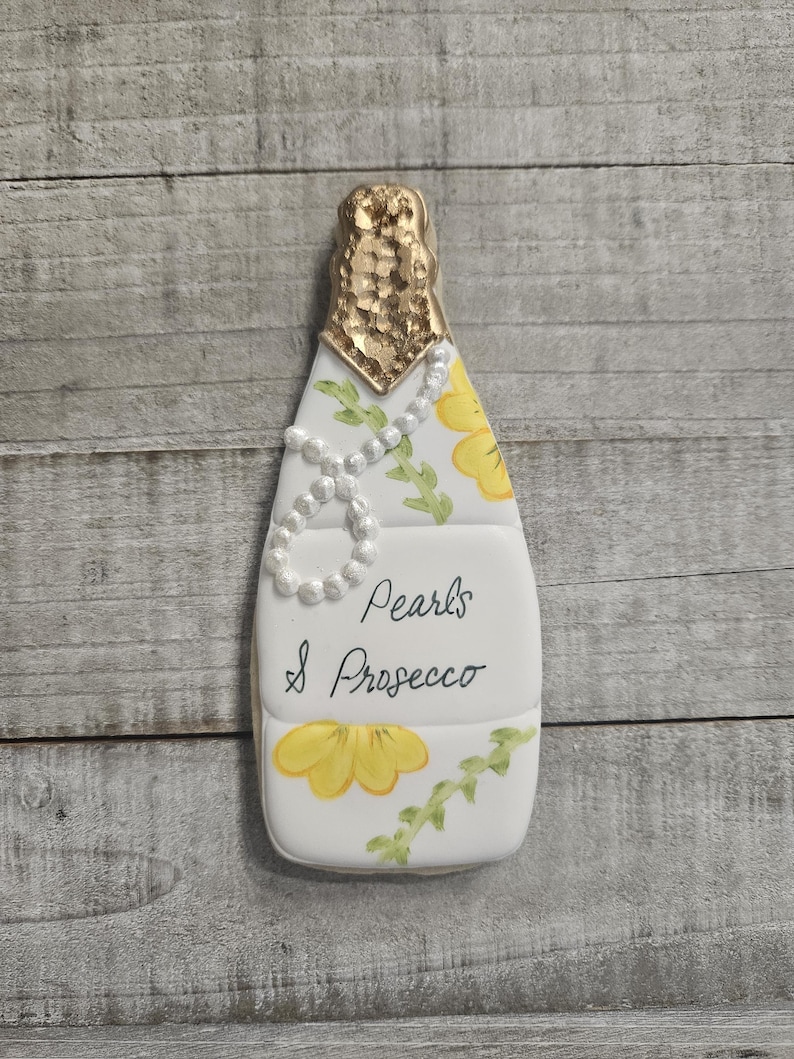 K&ouml;nnte beinhalten: Ein Champagnerflaschen-f&ouml;rmiger Keks mit goldenem Folienaufsatz, verziert mit einer Perlenkette und gelben Blumenakzenten. Die Worte "Pearls & Prosecco" sind auf der Vorderseite geschrieben.