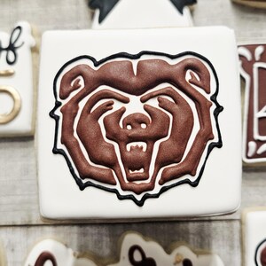 May include: A white cookie decorated with a brown bear head outline in black icing. The bear's face is brown with a white snout and black eyes.