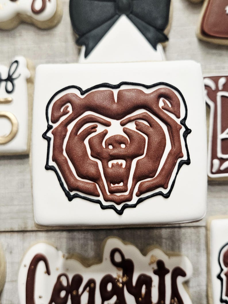May include: A white cookie decorated with a brown bear head outline in black icing. The bear's face is brown with a white snout and black eyes.