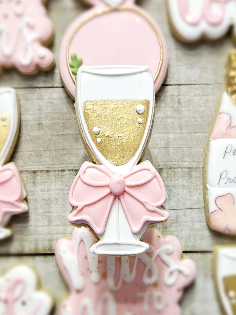 K&ouml;nnte beinhalten: Nahaufnahme von dekorierten Zuckerkeksen. Die Kekse haben die Form eines Sektglases, eines Rings und einer Sektflasche. Der Sektglaskeks hat eine rosa Schleife und goldfarbenen Sekt. Die Kekse sind mit wei&szlig;em und rosa Zuckerguss verziert.