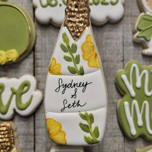 K&ouml;nnte beinhalten: Ein dekorierter Zuckerkeks in Form einer Champagnerflasche. Die Flasche ist wei&szlig; mit gelben Blumen und gr&uuml;nen Bl&auml;ttern. Der obere Teil ist goldfarben und der Keks tr&auml;gt die Aufschrift "Sydney & Seth". Das Datum "08.06.2025" ist ebenfalls vorhanden.