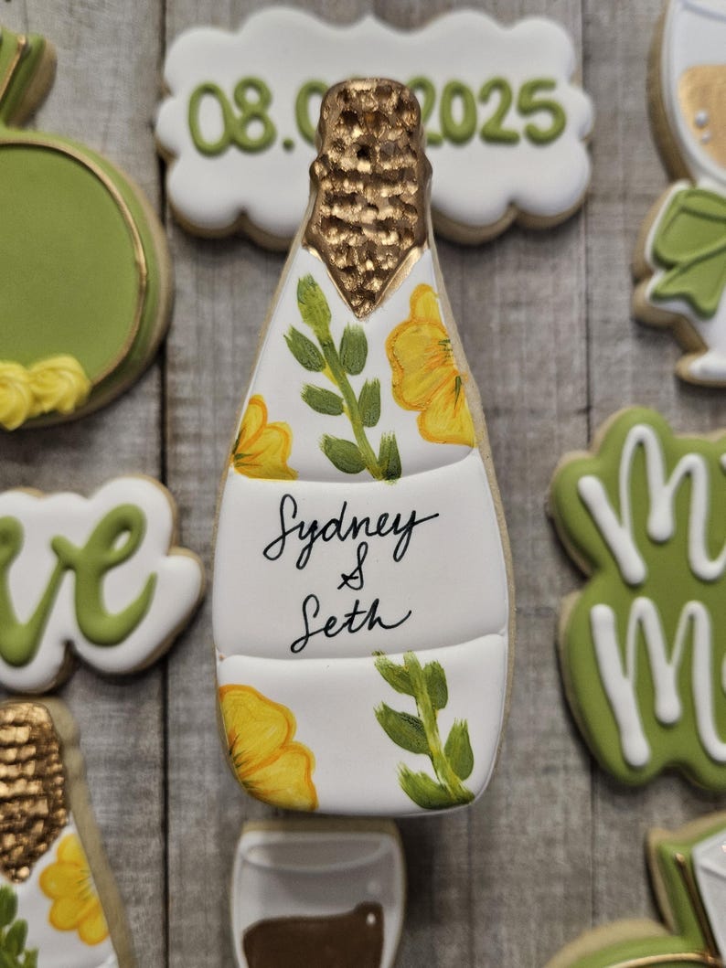 K&ouml;nnte beinhalten: Ein dekorierter Zuckerkeks in Form einer Champagnerflasche. Die Flasche ist wei&szlig; mit gelben Blumen und gr&uuml;nen Bl&auml;ttern. Der obere Teil ist goldfarben und der Keks tr&auml;gt die Aufschrift "Sydney & Seth". Das Datum "08.06.2025" ist ebenfalls vorhanden.