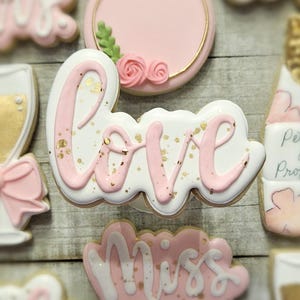 K&ouml;nnte beinhalten: Auswahl an dekorierten Keksen in Rosa und Wei&szlig;. Ein Keks schreibt "love" in rosa Zuckerguss mit Goldflocken. Weitere Kekse sind ein Ring, ein Sektglas und der Text "Miss to Mrs".
