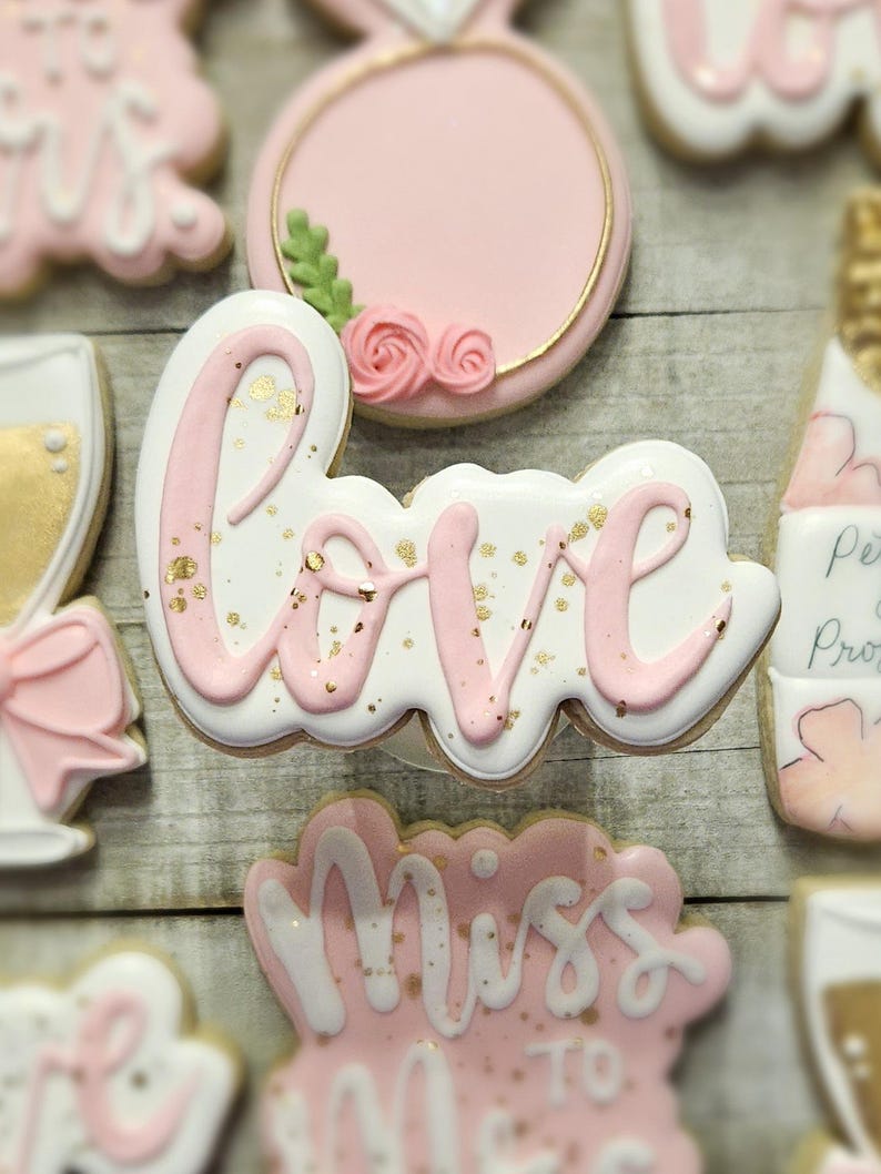 K&ouml;nnte beinhalten: Auswahl an dekorierten Keksen in Rosa und Wei&szlig;. Ein Keks schreibt "love" in rosa Zuckerguss mit Goldflocken. Weitere Kekse sind ein Ring, ein Sektglas und der Text "Miss to Mrs".