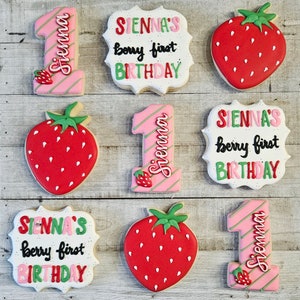 May include: Six decorated sugar cookies with pink and green icing. The cookies feature the number one, strawberries, and the text "SIENNA'S berry first BIRTHDAY".