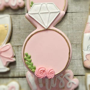 K&ouml;nnte beinhalten: Ein rosa Zuckerkeks in Form eines Diamant-Verlobungsrings. Der Ring hat einen goldenen Rand und ist mit einem wei&szlig;en Diamanten, gr&uuml;nen Bl&auml;ttern und rosa Rosen verziert. Weitere Kekse sind im Hintergrund zu sehen.