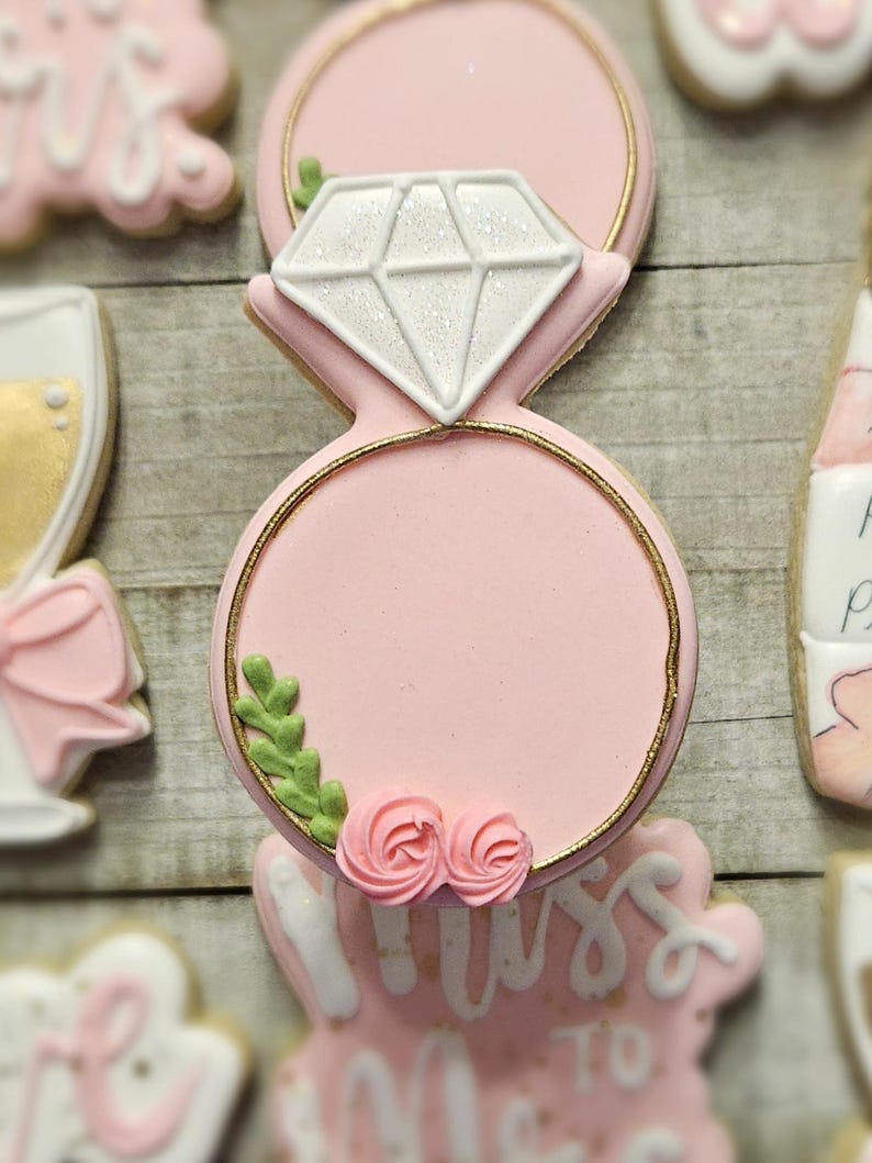 K&ouml;nnte beinhalten: Ein rosa Zuckerkeks in Form eines Diamant-Verlobungsrings. Der Ring hat einen goldenen Rand und ist mit einem wei&szlig;en Diamanten, gr&uuml;nen Bl&auml;ttern und rosa Rosen verziert. Weitere Kekse sind im Hintergrund zu sehen.