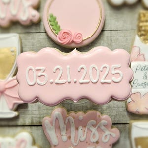 K&ouml;nnte beinhalten: Rosa glasierte Kekse mit wei&szlig;em Zuckerguss. Ein Keks tr&auml;gt das Datum 03.21.2025. Weitere Kekse zeigen einen Ring, ein Sektglas und den Schriftzug "Miss to Mrs."