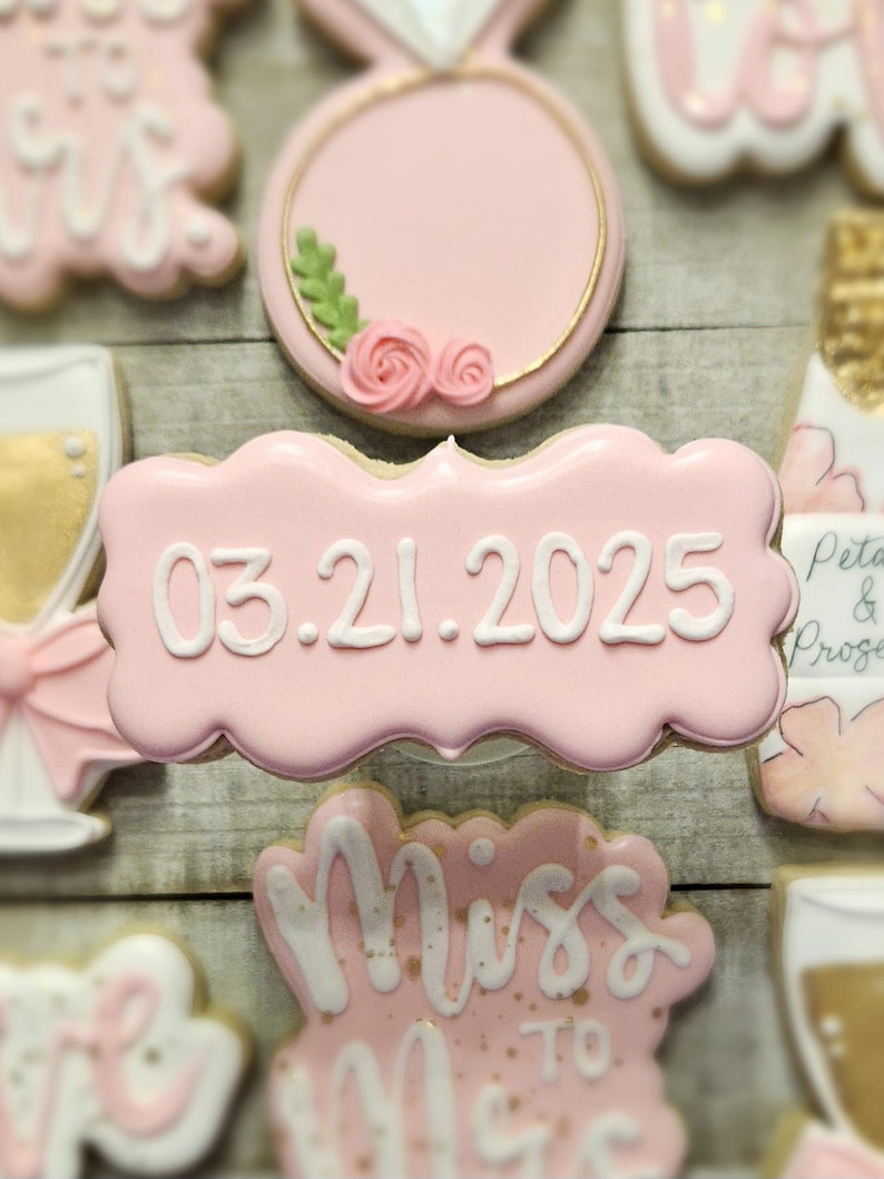 K&ouml;nnte beinhalten: Rosa glasierte Kekse mit wei&szlig;em Zuckerguss. Ein Keks tr&auml;gt das Datum 03.21.2025. Weitere Kekse zeigen einen Ring, ein Sektglas und den Schriftzug "Miss to Mrs."