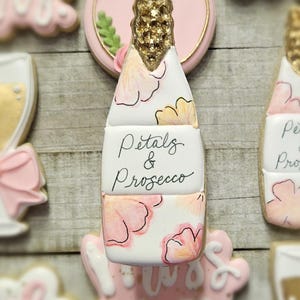 K&ouml;nnte beinhalten: Ein Keks in Champagnerflaschenform mit goldenem Glitzer auf der Oberseite und rosa Blumendesigns. Der Keks tr&auml;gt die Aufschrift "Petals & Prosecco". Weitere Kekse im Hintergrund sind mit rosa Zuckerguss und goldenen Akzenten verziert.