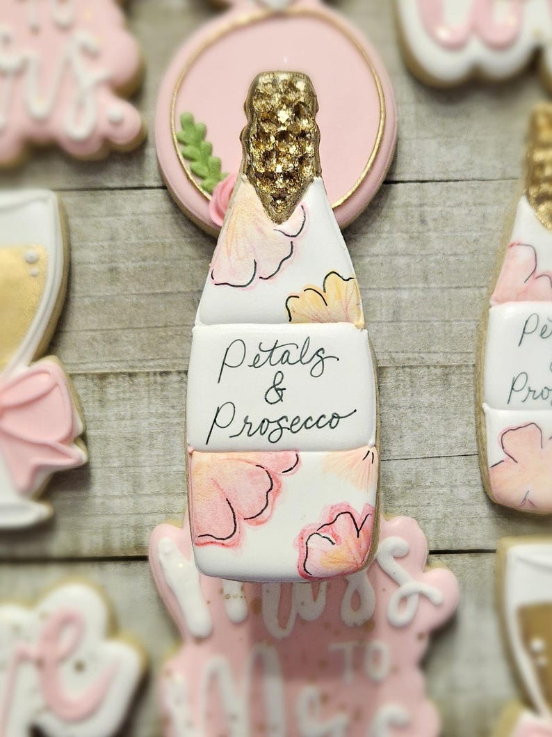 K&ouml;nnte beinhalten: Ein Keks in Champagnerflaschenform mit goldenem Glitzer auf der Oberseite und rosa Blumendesigns. Der Keks tr&auml;gt die Aufschrift "Petals & Prosecco". Weitere Kekse im Hintergrund sind mit rosa Zuckerguss und goldenen Akzenten verziert.