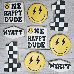 May include: Nine decorated sugar cookies with black and white icing. The cookies feature the words "One Happy Dude", "Wyatt", and the number "1". Three of the cookies feature a yellow smiley face with lightning bolts for eyes.