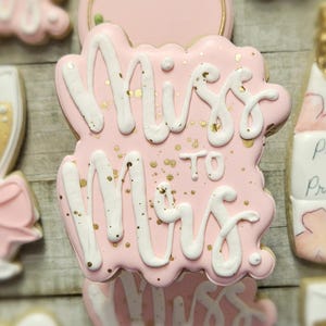 K&ouml;nnte beinhalten: Rosa glasierter Keks mit wei&szlig;em Schreibschrift-Text "Miss to Mrs." und Goldflocken. Weitere Kekse im Hintergrund sind eine Champagnerflasche und ein Champagnerglas. Die Kekse sind wahrscheinlich f&uuml;r eine Brautparty.