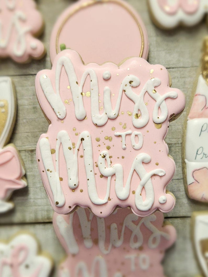 K&ouml;nnte beinhalten: Rosa glasierter Keks mit wei&szlig;em Schreibschrift-Text "Miss to Mrs." und Goldflocken. Weitere Kekse im Hintergrund sind eine Champagnerflasche und ein Champagnerglas. Die Kekse sind wahrscheinlich f&uuml;r eine Brautparty.