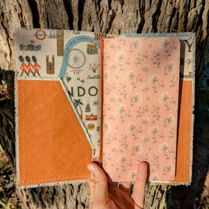 May include: A fabric notebook cover featuring a London map print with illustrations of the London Eye and guards, paired with a tan faux leather pocket. The interior contains a notebook with a light pink cover and a white floral pattern.