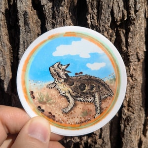 May include: A round sticker featuring a detailed illustration of a horned lizard in a desert scene with ants, blue sky, and clouds. The sticker has an orange and white border, and the artist's signature is visible.
