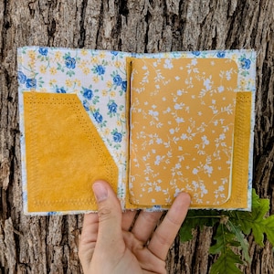 May include: Open journal with a floral pattern. The cover is white with blue flowers, and the interior pages are yellow with white flowers. The journal has a mustard-colored pocket on the left side.