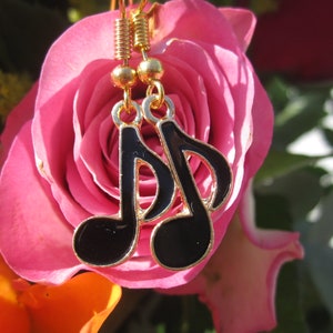 Könnte beinhalten: Goldfarbene Ohrringe mit schwarzen Noten. Die Ohrringe hängen an goldfarbenen Haken, vor dem Hintergrund einer rosa Rose und anderer bunter Blumen. Diese Ohrringe sind ein Accessoire zum Thema Musik.
