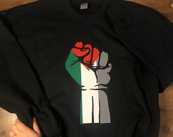 May include: Black sweatshirt featuring a raised fist design in red, green, white, and gray. The graphic is a stylized representation of a fist with the colors of a flag. The sweatshirt is made of a soft material.