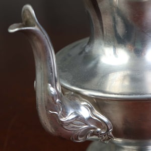 May include: A silver teapot with an ornate, floral design on the spout. The teapot has a polished, reflective surface.