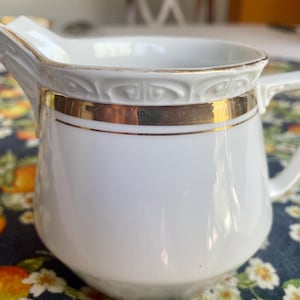 May include: White porcelain creamer with a gold band and trim. The creamer has a handle and a spout for pouring. The design includes a decorative pattern around the rim. The creamer is likely vintage or antique.