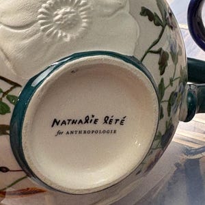 May include: A white ceramic mug with a floral design and a teal rim. The mug features the text "Nathalie Lete" and "for Anthropologie" in black script. The mug has a handle and is decorated with embossed floral details.