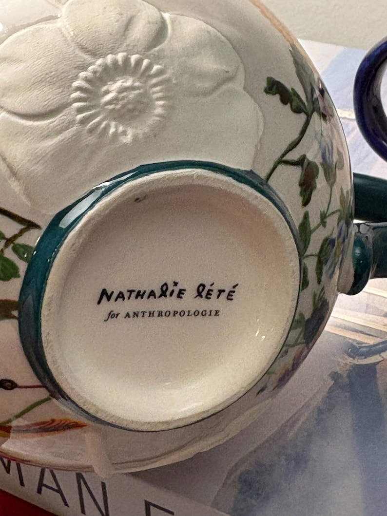 May include: A white ceramic mug with a floral design and a teal rim. The mug features the text "Nathalie Lete" and "for Anthropologie" in black script. The mug has a handle and is decorated with embossed floral details.