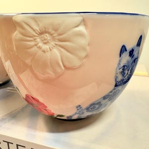 May include: A pink ceramic bowl with a blue rim and a white embossed flower. The bowl features a blue and white cat illustration and a red flower. The bowl is likely for serving or display.