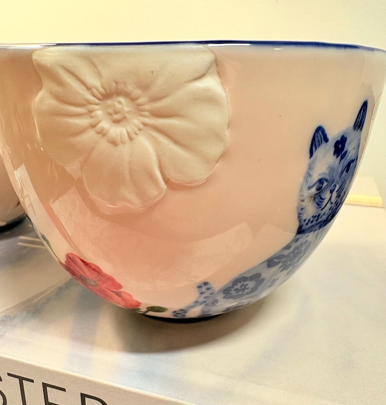 May include: A pink ceramic bowl with a blue rim and a white embossed flower. The bowl features a blue and white cat illustration and a red flower. The bowl is likely for serving or display.