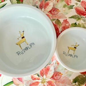 May include: Two white ceramic bowls with a hand-painted illustration of a yellow reindeer with the word "Rudolph" below. The bowls are on a floral patterned surface with red and white flowers.