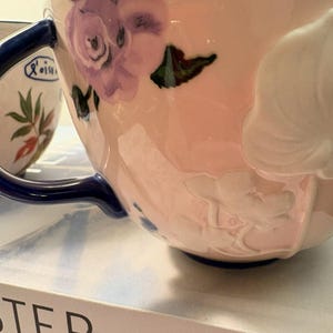 May include: Close-up of a pink ceramic teacup with a blue handle and rim. The cup is decorated with purple floral designs and embossed white flowers. A second cup with a floral design is partially visible in the background.