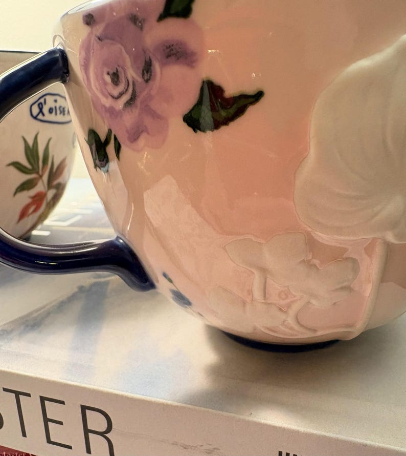 May include: Close-up of a pink ceramic teacup with a blue handle and rim. The cup is decorated with purple floral designs and embossed white flowers. A second cup with a floral design is partially visible in the background.