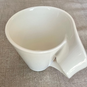 May include: A white ceramic coffee cup with a unique handle design. The cup has a wide opening and a curved handle that extends from the side. The cup is made of a glossy, smooth material.