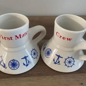 May include: Two white ceramic mugs with blue and red nautical designs. One mug says "First Mate" and the other says "Crew".