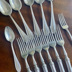 Puede incluir: Un juego de cucharas y tenedores de plata con mangos adornados. Las cucharas tienen un cuenco redondeado y los tenedores tienen cuatro dientes.