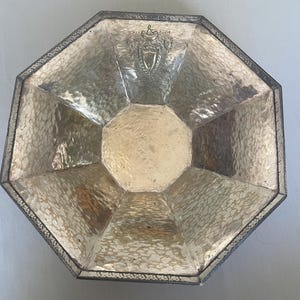 May include: An octagonal silver-toned serving dish with a hammered texture. The dish has a decorative border and a crest design in the center. The dish is likely vintage and suitable for serving appetizers or small dishes.
