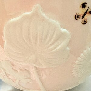 May include: A pink ceramic bowl with a raised floral design and a small butterfly. The bowl has a blue rim and a black base. The floral design is white and the butterfly has black and brown details.