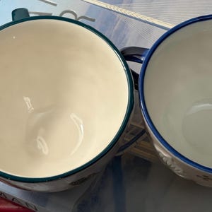 May include: Two ceramic soup mugs. One mug has a teal rim and handle, the other has a blue rim and handle. Both mugs have a white interior. The mugs are sitting on a book.