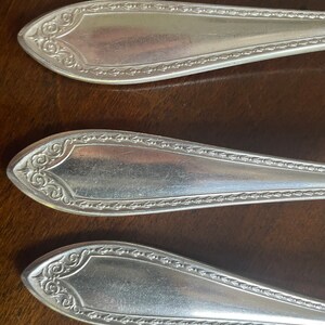 May include: Three silver-plated spoons with ornate handles and a beaded edge. The spoons are arranged in a row on a wooden surface.