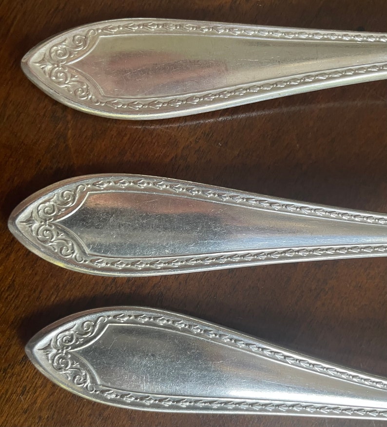 May include: Three silver-plated spoons with ornate handles and a beaded edge. The spoons are arranged in a row on a wooden surface.