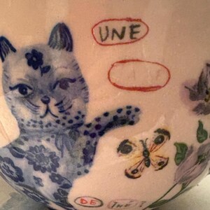 May include: A ceramic teacup with a light pink background, featuring a blue cat illustration with floral details. A yellow butterfly and the text "UNE" and "DE THE?" are also visible.