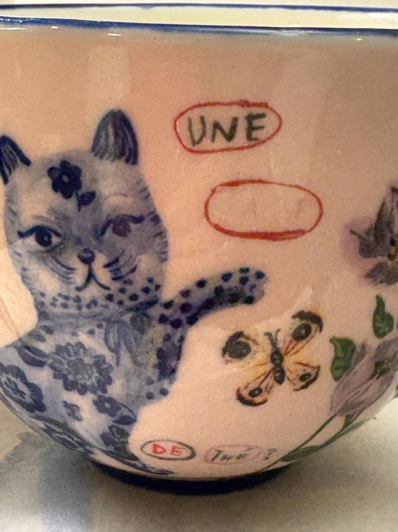 May include: A ceramic teacup with a light pink background, featuring a blue cat illustration with floral details. A yellow butterfly and the text "UNE" and "DE THE?" are also visible.