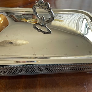 May include: A rectangular silver-toned serving dish with a removable lid featuring a decorative handle. The base has an intricate lattice-style openwork border, two side handles, and four small feet. The polished surface reflects the surroundings.